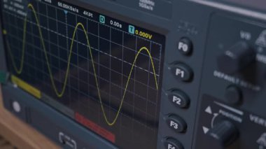 Dijital osiloskop ekranında sarı bir sinüs dalgası elektrik sinyali var. Kamera elektronik test ekipmanlarını tarıyor. Veri analizi, sinyal işleme ve araştırma kavramı.