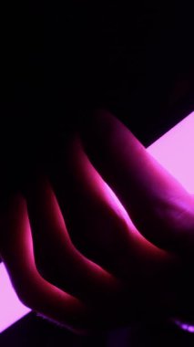 Bir el, karanlıkta parıldayan mor neon bir ışık tüpünü kavrayıp çarpıcı ışık sayesinde deri dokusunu ortaya çıkarıyor. Siber estetik, gece hayatı, gizem ve sanatsal yaratıcılık kavramı.