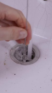 Bir el, kirli beyaz seramik banyo lavabosundaki tıkanıklığı temizlemek için metal bir tel kullanıyor. Ev bakımı, tesisat sorunları ve DIY onarım çözümleri kavramı.