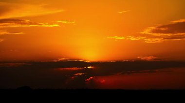 Karanlık bir bulut katmanının ardında batan altın güneşin zamanı gökyüzünde canlı turuncu bir parıltı bırakıyor. Ufuk karanlık bir siluet olarak kalır dramatik bir manzara yaratır..