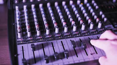 Ses seviyelerini kontrol etmek için bir fünyeyi kaydırarak profesyonel bir ses konsolunu elle çalıştırmak. Sahnede bir stüdyoda veya canlı yayında dramatik kırmızı, mor ve mavi yanıp sönen ışıklar yer alıyor..