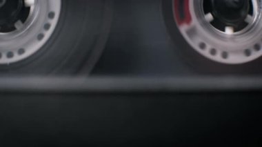 Makro görünüm, şeffaf ses kasetlerinin teypte döndüğünü ortaya çıkarmak için yukarı doğru eğiliyor. Bu çekim, arşivlenmiş sorgulama ya da polis delillerinin toplanmasını çağrıştırıyor..