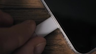 Beyaz bir USB-C kablosunu ahşap bir masada şarj eden bir akıllı telefondan ayıran parmakların makro görüntüsü. Şeffaf vaka ve siyah ekran modern mobil teknolojiyi vurguluyor.