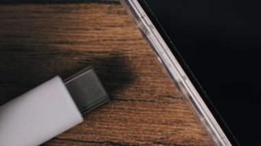 Bir elin makro görüntüsü, cep telefonu güç portuna beyaz bir kablo yerleştiriyor. Koyu ekran şeffaf kabuklu cihaz desenli ahşap bir arka planda duruyor..