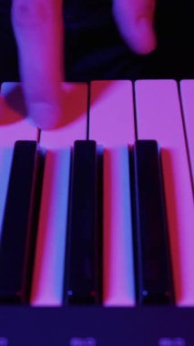 Müzisyen elleri bir sentezleyici klavyesinde melodi çalar. Dramatik pembe ve mavi neon ışıklar karanlık sahneyi aydınlatıyor..