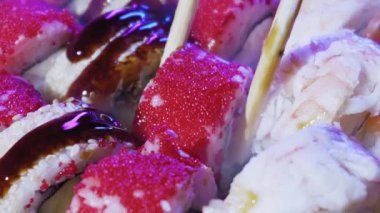 Tahta çubuklar kalabalık bir tabaktan kırmızı balık yumurtası ile kaplı bir suşi rulosu alır. Serin mor ışık, pirinç ve kremalı dolgunun dokusunu vurgular..