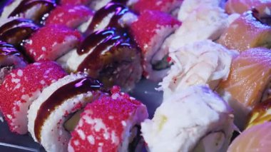 Ahşap yemek çubukları kırmızı yumurtayla kaplanmış suşi rulosunu ve tabaktan koyu sos kaldırır. Parlak yapay ışıklandırma balık yumurtası ve kremalı malzemelerin dokusunu vurgular..