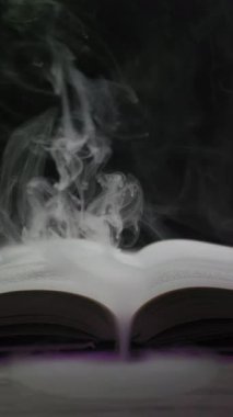 Açık bir kitap, koyu bir arkaplana doğru yükselen kalın ve mistik bir duman yayar. Işığın rengi mora döndüğünde. Sihir, fantezi, hikaye anlatma ve yasak büyüler kavramı..