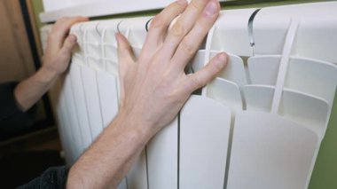 Bir adamın elleri, odadaki sıcaklığı ve sıcaklığı ölçmek için beyaz bir ısıtıcı kalorifere dokunuyor. Isınma mevsimi kavramı, ev konforu, yükselen enerji fiyatları ve ısı tasarrufu.