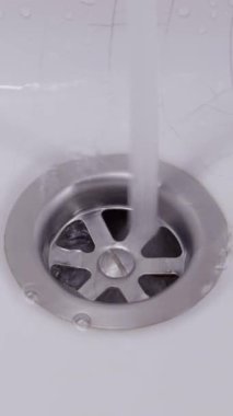 Sabit bir berrak su akışı paslanmaz çelik lavabo giderine akar, girdaplı kabarcıklar oluşturur ve beyaz leğende su sıçratır. Ev hijyeni, tesisat ve atık yönetimi kavramı.