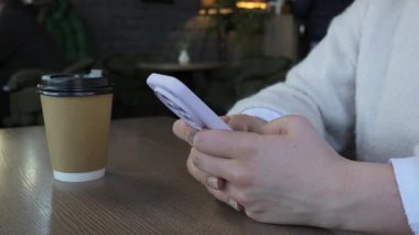 Kağıt kahve fincanının yanındaki ahşap bir kafe masasında otururken açık mor bir akıllı telefonu tutan ve daktilo eden kadın ellerini kapatın. Bu modern bir yaşam tarzı konseptini temsil ediyor..