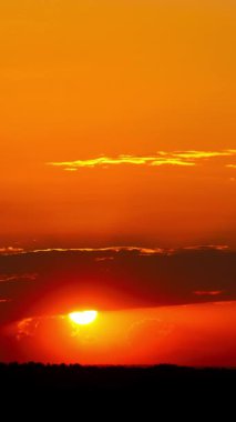 Karanlık bulutların arkasından süzülen parlak güneşin dikey zaman çizelgesi ve parlak turuncu renklerle gökyüzünü boyuyor. Aşağıdaki manzara hala simsiyah bir siluet..