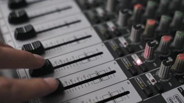 Profesyonel karıştırma konsolunda iki fader seviyesini ayarlayan bir ses mühendisinin yakın plan açılı çekimi. Panelde beyaz kanal şeritleri ve stüdyo ortamında kırmızı ışık görünüyor..