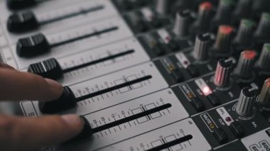 Bir ses yapımcısının parmaklarının makro çekimi profesyonel bir ses konsolunda çoklu fadorları aşağı yukarı hareket ettiriyor. Ses düzeyi bu sığ odak çekiminde görünür desibel ölçeğinde ayarlanır..