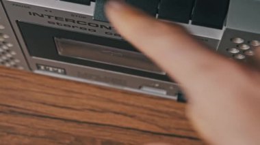 Gümüş bir stereo çalardaki fırlatma tuşuna basan bir elin yukarıdan görünüşü. Kullanıcı, teybi kapatmadan önce sarı etiketli eski bir kaseti kaldırır.