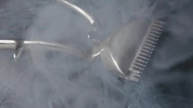 Klasik manuel saç makası koyu, yansıtıcı bir yüzeyde yatar. Ruhsal beyaz duman yavaşça süzülür ve klasik berber aletinin etrafında döner, gizemli bir atmosfer yaratır..