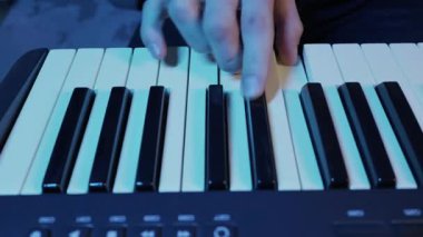 Erkek eller elektronik piyano klavyesinde bir melodi çalar. Sahne parlak mavi ve morumsu neon ışıklarla aydınlatılıyor, modern ve yaratıcı bir hava yaratıyor..