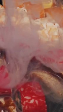 Somon, yılan balığı ve tobiko ile çeşit çeşit suşi ruloları kalın beyaz bir duman tabakasının altında oturur. Soğuk tazeliği vurgulayan taze Japon deniz ürünleri düzenlemesi üzerine ağır kuru buz buharı akar..