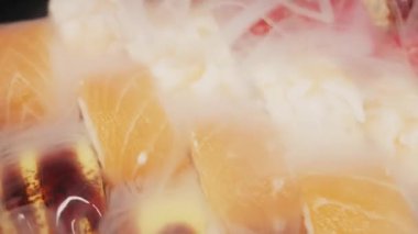 Somon ve kırmızı tobiko ile kaplanmış çeşitli suşi dürümleri siyah bir tabakta oturur. Yoğun beyaz sıvı nitrojen dumanı Japon mutfağının etrafında dramatik bir sunumla dönüyor..