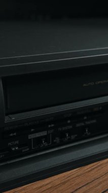 Erkek eli siyah VHS bandını eski video kaset kaydedicinin mekanik yuvasına itiyor. Sinematik ışıklandırma, klasik elektronik yüzey üzerindeki toz dokusunu vurguluyor.