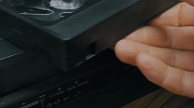 Erkek el, siyah VHS kaset kasetini retro video oynatıcının yuvasına yerleştirir. Mekanik yükleme sistemi bant ahşap masa yüzeyinde cihazın içine çeker.