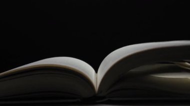 Açık bir kitap karanlık bir odada duruyor. Sayfalar yavaşça dönüyor ve çırpınıyor. Tek bir ışık kaynağıyla aydınlanıyor. Hikaye anlatma ve bilgi kavramı.