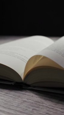 Ahşap bir yüzey üzerine açık bir kitabın dikey videosu. Sayfalar yavaşça döner ve hafif bir esintide karanlık bir arka plana doğru çırpınır. Bu kavram okuma ve bilgiyi temsil eder..