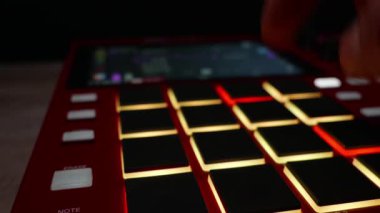 Müzisyenlerin parmakları kırmızı bir davul makinesinin ışıklı pedlerine dokunuyor. Elektronik müzik üretimi, yaratıcı ses mühendisliği ve profesyonel stüdyo çalışma akışı kavramı.
