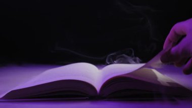 Bir el, karanlıkta dönen sis ve mor ışıkla çevrili açık bir kitabın sayfasını çevirir. Büyü kavramı, gizemli hikaye anlatımı, fantezi dünyası, ve antik bilgi.