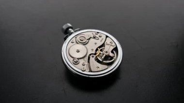 Hızlandırılmış bir makro zaman atlaması siyah bir masada hızla dönen antika bir saat mekanizmasının dişlilerini ortaya çıkarır. Son teslim tarihi baskısı, hassas mühendislik ve zamanın kısacık doğası kavramı..