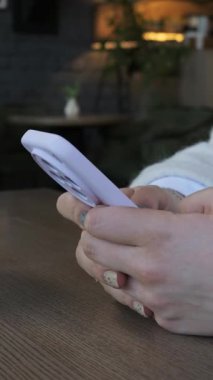 Ahşap bir kafe masasında duran bir kadının elleri mor bir akıllı telefonu tutuyor ve daktilo ediyor. Bulanık arka plan, kafenin içini gösteriyor. Bu modern bir iletişim konseptini gösteriyor..