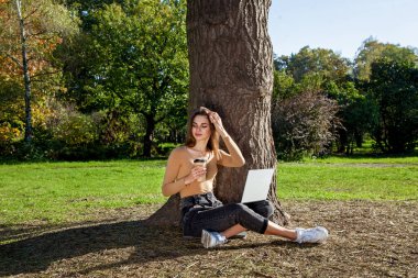 Bir kız elinde bir fincan kahveyle parkta bir ağacın yanında oturuyor ve dizüstü bilgisayarla çalışıyor. Açık havada serbest çalışmak. Covid 19 salgınından dolayı ofis dışında çalışıyor.