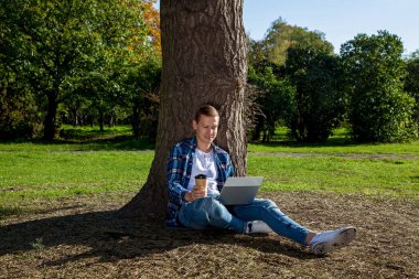 Adam elinde bir fincan kahveyle parkta bir ağacın yanında oturuyor ve dizüstü bilgisayarla çalışıyor. Açık havada serbest çalışmak. Covid 19 salgınından dolayı ofis dışında çalışıyor.