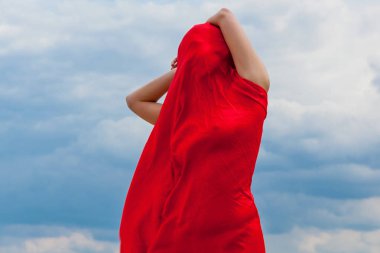 Kumda kırmızı bir bez giymiş bir kız bulutlu bir gökyüzünün arkaplanına karşı bir fotoğrafçıya poz veriyor. Rüzgardaki kırmızı kumaş kızın vücuduna sarılıyor..