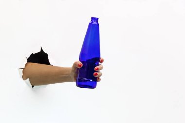 Kırmızı manikürlü, yırtık beyaz kağıtlı bir kadın elinde mavi bir bira şişesi tutuyor. Yırtık beyaz kağıtlar arasında