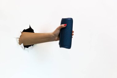 Kırmızı tırnak cilasıyla yırtılmış beyaz kağıdı delen bir kadın eli kablosuz taşınabilir bir hoparlör tutuyor. Yırtık beyaz kağıtlar arasında