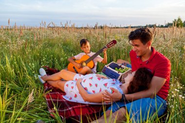 Genç ebeveynler ve oğulları güneşli bir günde buğday tarlasında piknik yapıyorlar. Oğlu ailesi için gitar çalıyor..