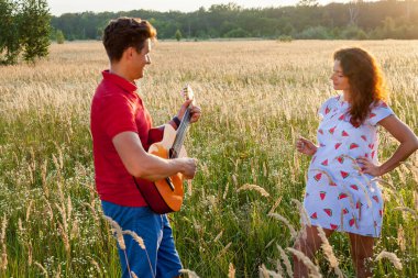 Gitarlı genç bir adam güneşli bir günde hamile karısının yanında duruyor..