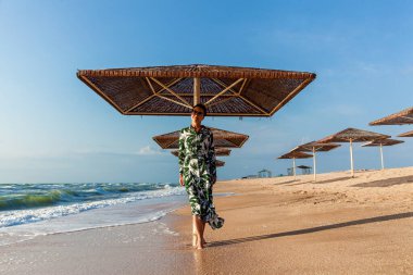 Plajda güneş şemsiyesinin yanında yeşil yapraklı beyaz elbiseli kız poz veriyor. Sahilde poz veren elbiseli bir kız.