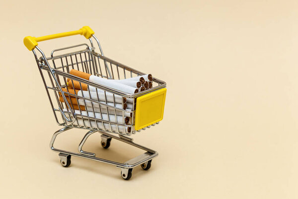 Metal shopping cart with cigarettes on a beige background. Concept objects for supermarket. Photo with place for your text and design.