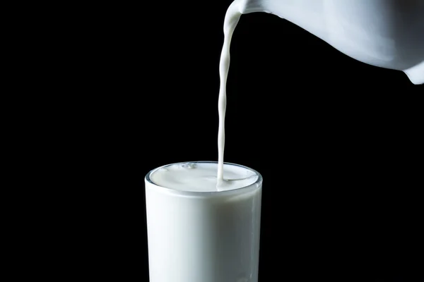 O fluxo derrama o leite de um jarro em um copo isolado em um fundo preto — Fotografia de Stock