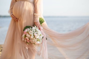Beautiful pregnant woman with flowers holds hands on belly in nature. Young woman in maternity dress waiting for baby birth. Pregnancy, Motherhood, Mothers Day Holiday concept.