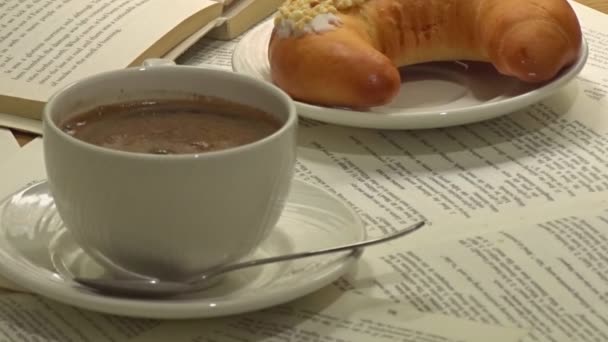 A white cup of really hot smoking coffee on sheets of book papers with dessert and open book on background — Stock Video