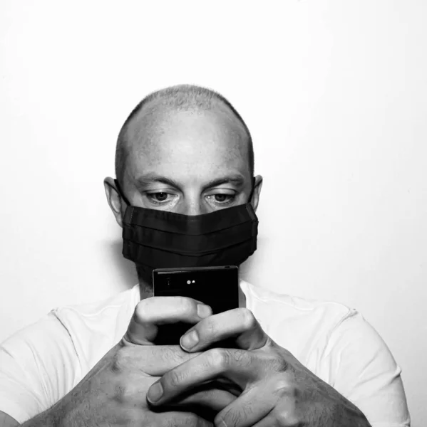 Black and white image of man wearing face mask for virus protection, reading news on his mobile phone while in isolation. . High quality photo