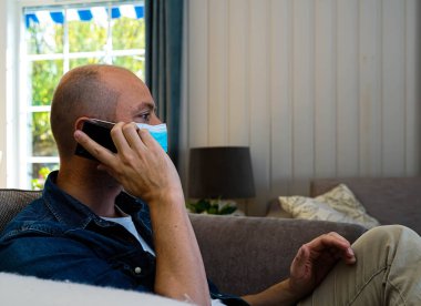 Man wearing a face mask sitting in his home talking on the phone. . High quality photo
