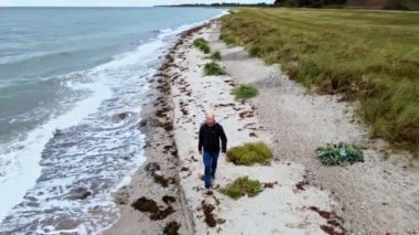 Sonbaharda kumlu bir sahilde kameraya doğru yürüyen bir adam.