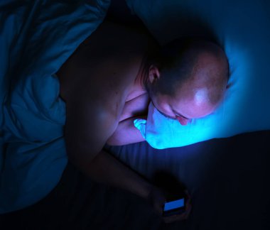 Geceleri yatağında telefonunu kullanan ve yatak odasını aydınlatan zararlı mavi ışığı olan bir adam. Yüksek kalite fotoğraf