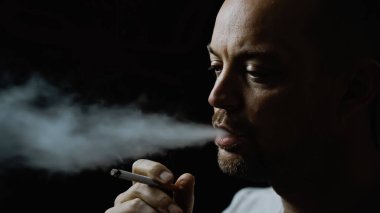 Sigara içen ve ağzından duman çıkaran bir adam. Siyah arkaplanda sığ bir alan derinliğinde izole edilmiş. Yüksek kalite fotoğraf