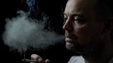 Sigara içen ve ağzından duman çıkaran bir adam. Siyah arkaplanda sığ bir alan derinliğinde izole edilmiş. Yüksek kalite fotoğraf