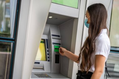 Koruyucu maskeli iş kadını para çekmek için ATM kullanıyor. Covid-19 virüs salgını konsepti.